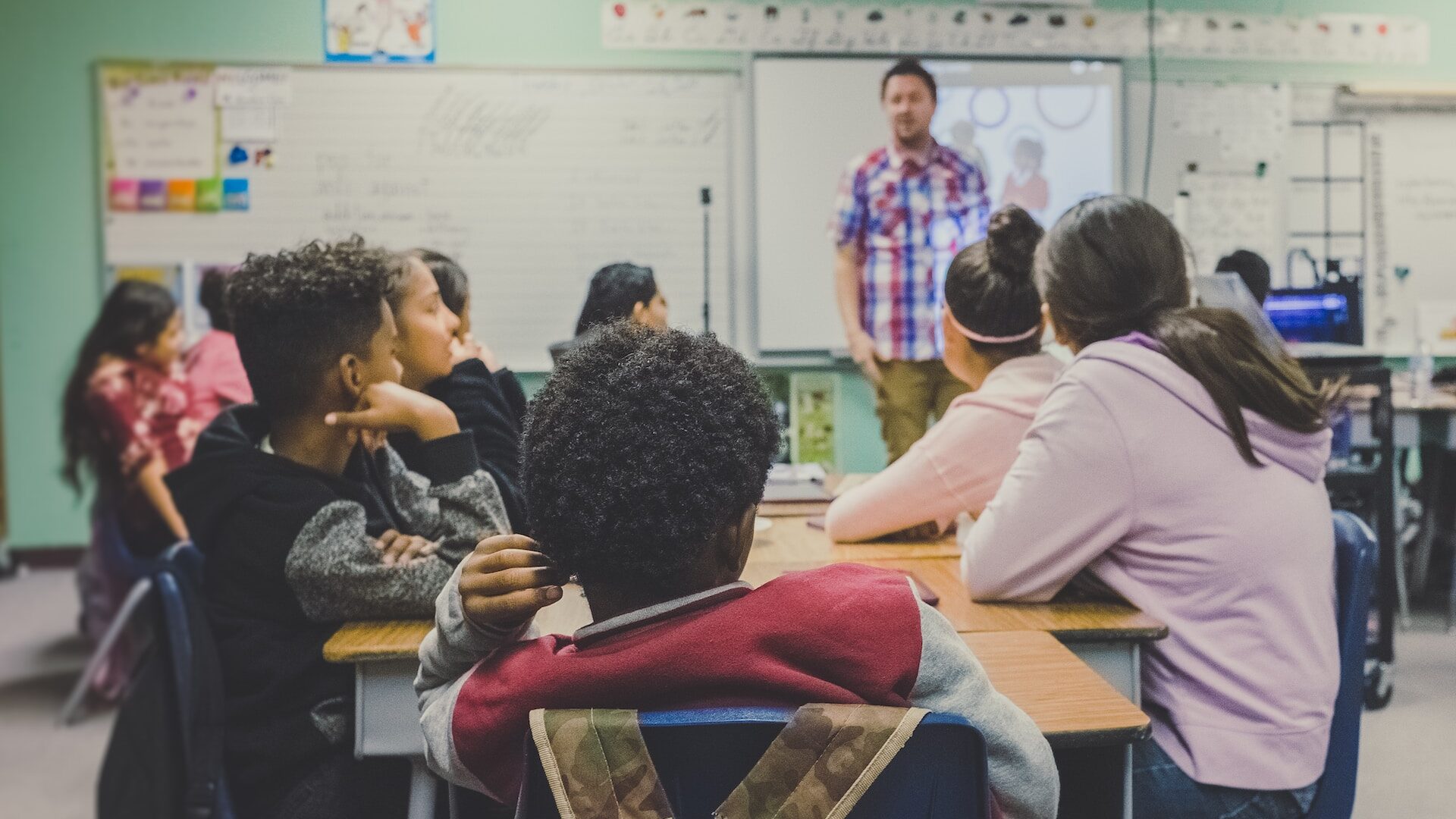 A group of children sit in a classroom with a teacher stood at the front of the class.