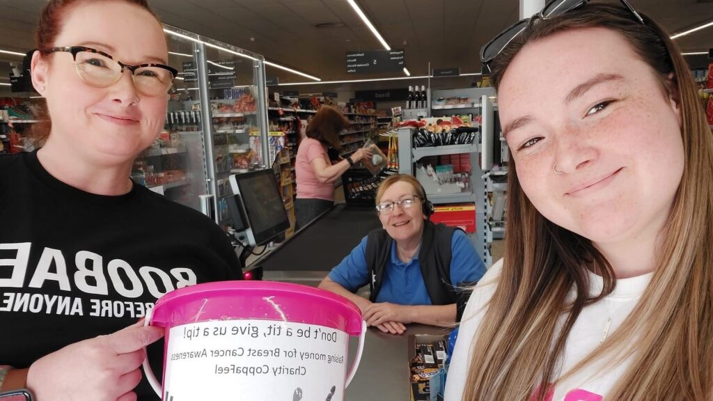 Coronation Champion Emilia McRobie and another student holding a collection bucket smile at the camera while standing in front of a supermarket checkout and member of staff.
