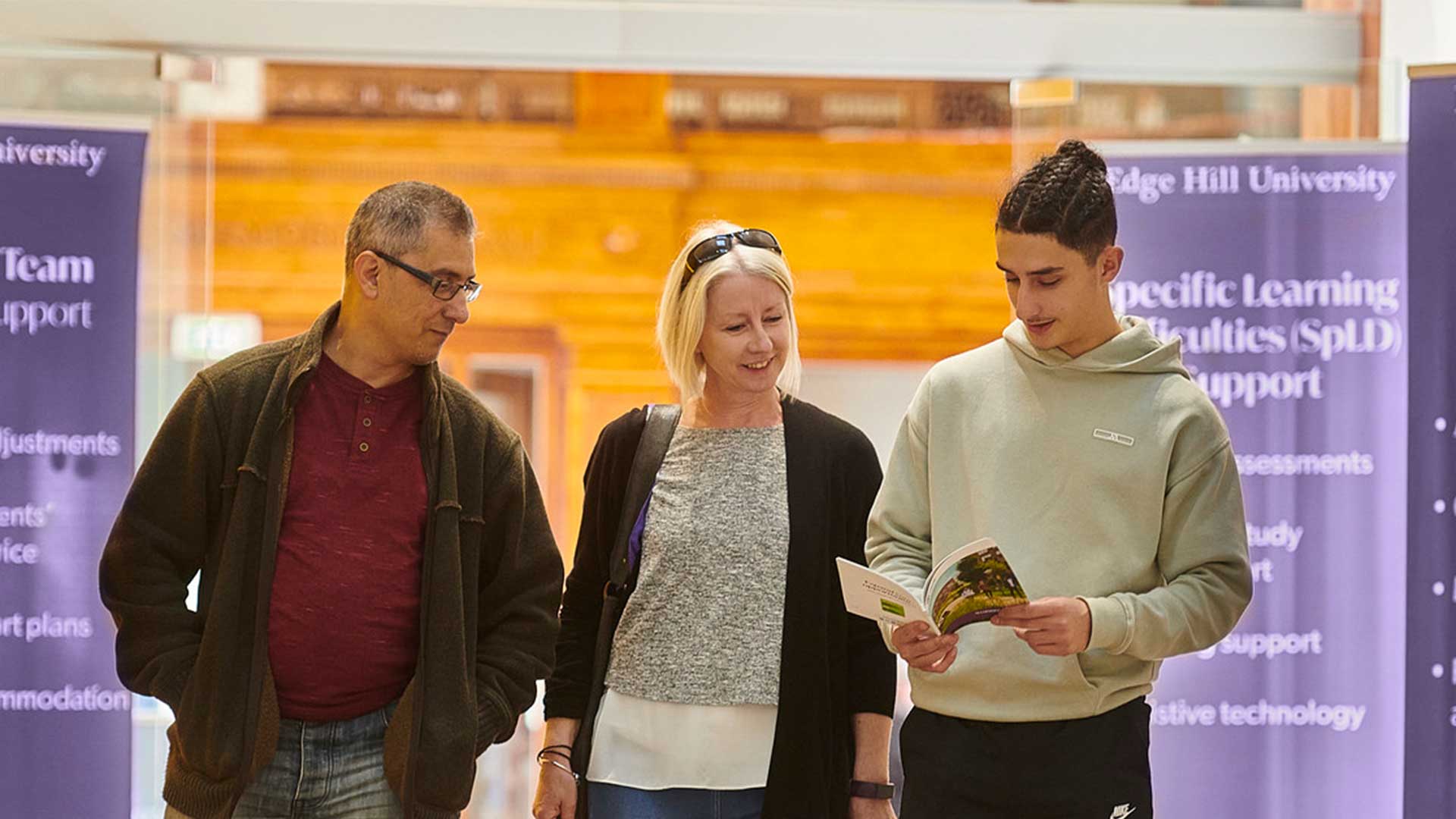A student walks through the Hub with their parents on an Open Day. They are looking through a printed Edge Hill University guide