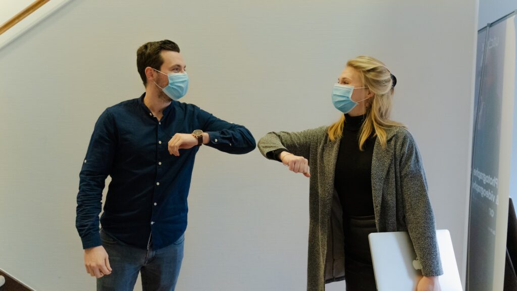 Two people wearing masks touch elbows.