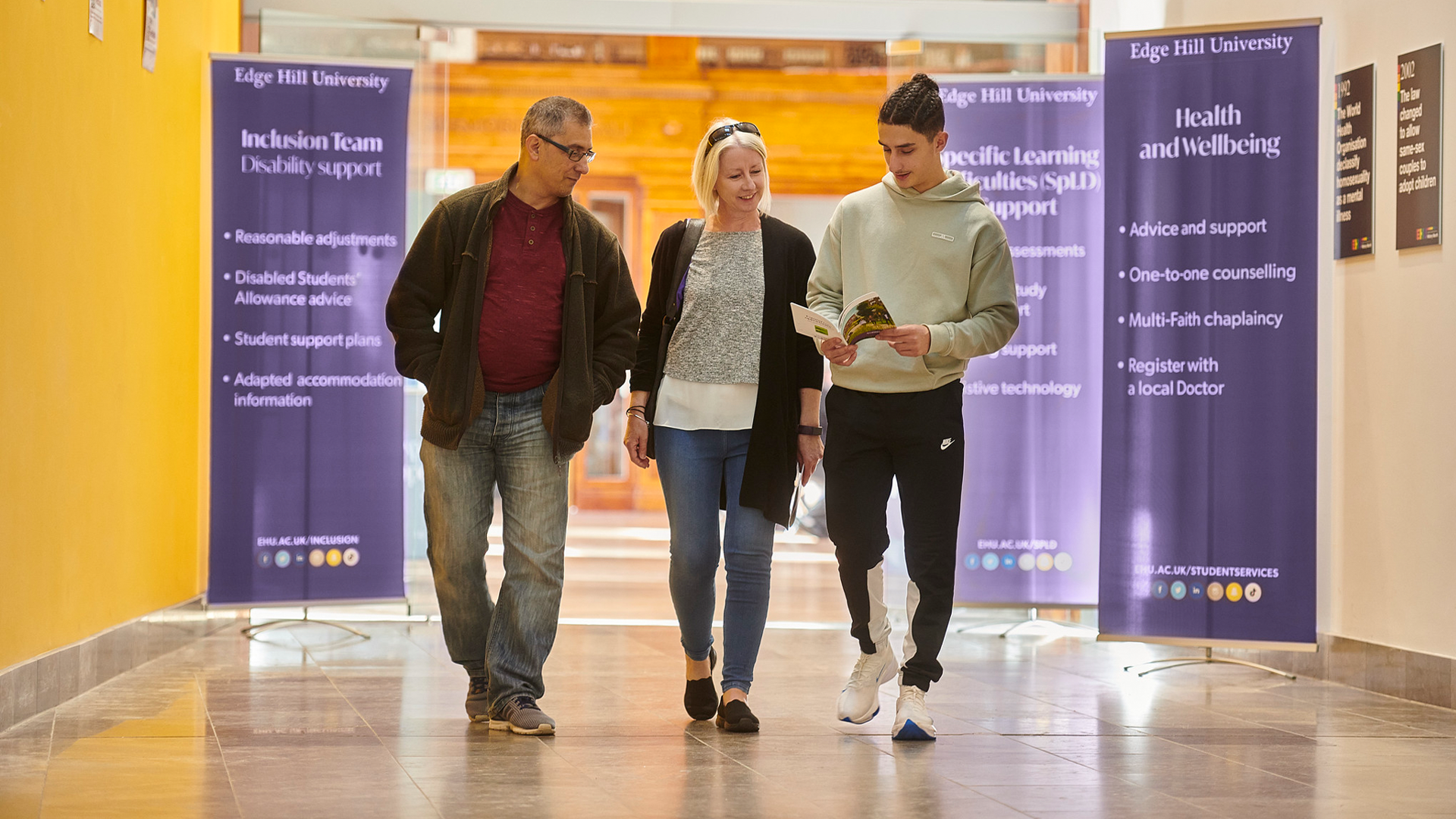 Student and their parents walk through Hale Hall on an open day. Signs advertising Health and Wellbeing and the Inclusion Team can be seen in the background.