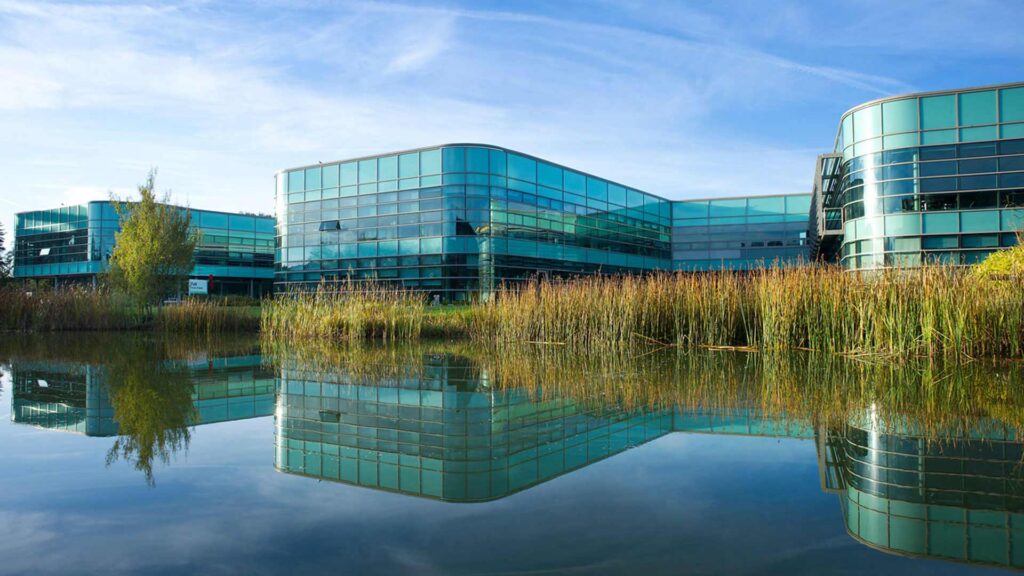Exterior of the Faculty of Health Social Care and Medicine building. The reflection of the building is on the surface of the lake with the lake reeds framing the building.