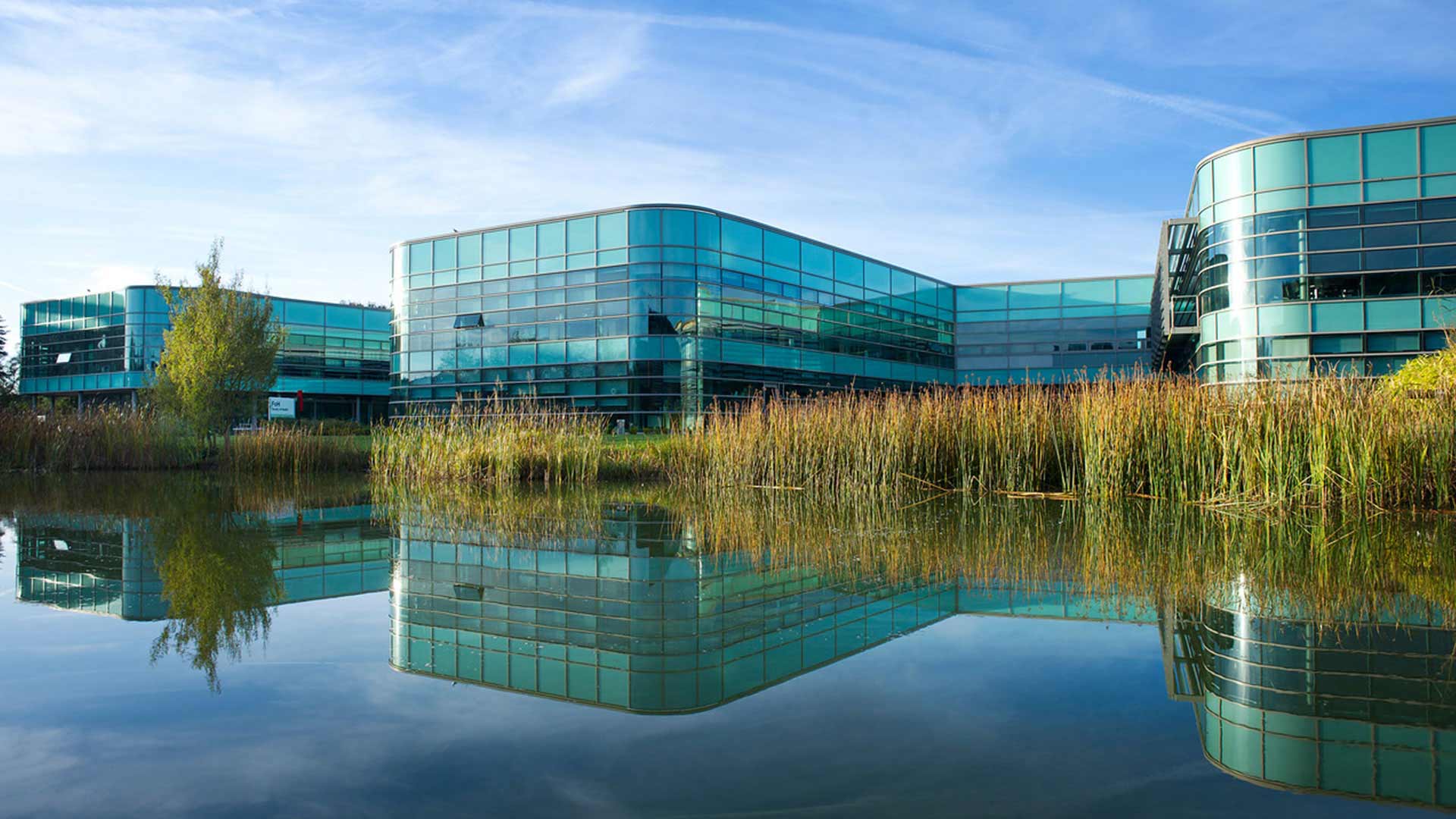 Exterior of the Faculty of Health, Social Care and Medicine building. The reflection of the building is on the surface of the lake with the lake reeds framing the building.