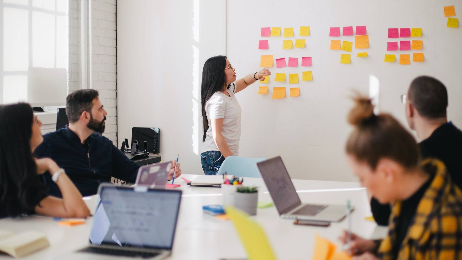 Members of an office team are sat around a large table with laptops. A member of the team is stood at the front of the room arranging sticky notes on a wall.