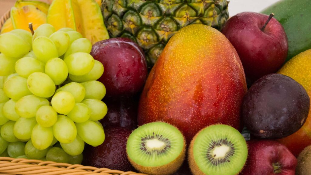 A close up shot of a basket of fruit. It includes grapes, kiwis, apples, plums, a mango, a pineapple and bananas.