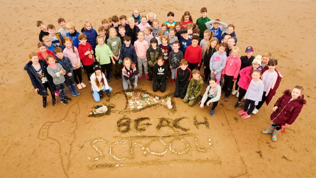 A group of children standing on a beach with the words Beach School written in the sand.