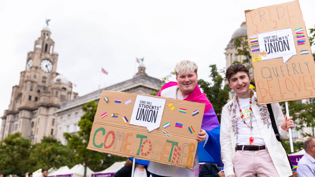 Jack and Ben, 2022/23 Presidents at LCR Pride march holding signs. Sign 1 says: 'Come together.' Sign 2 says 'Pride 2022! Queerly Be Loved' Photo by Students' Union