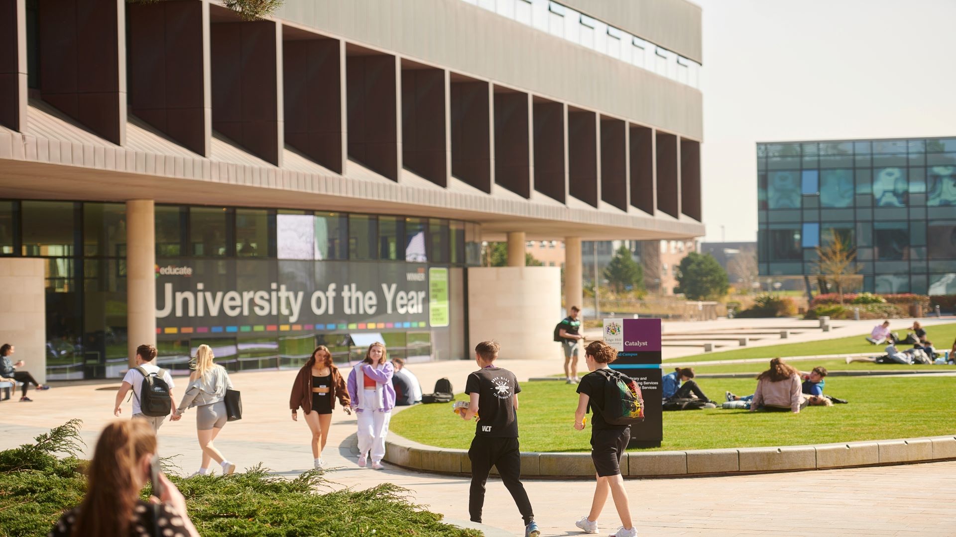 Students take a walk and sit on the grass outside the Catalyst. Catalyst has 'University of the Year' on the side of it.