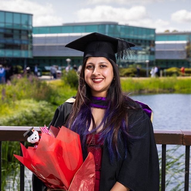 Isha Bhukya at graduation in their gown and hat with a bouquet of flowers in hand.
