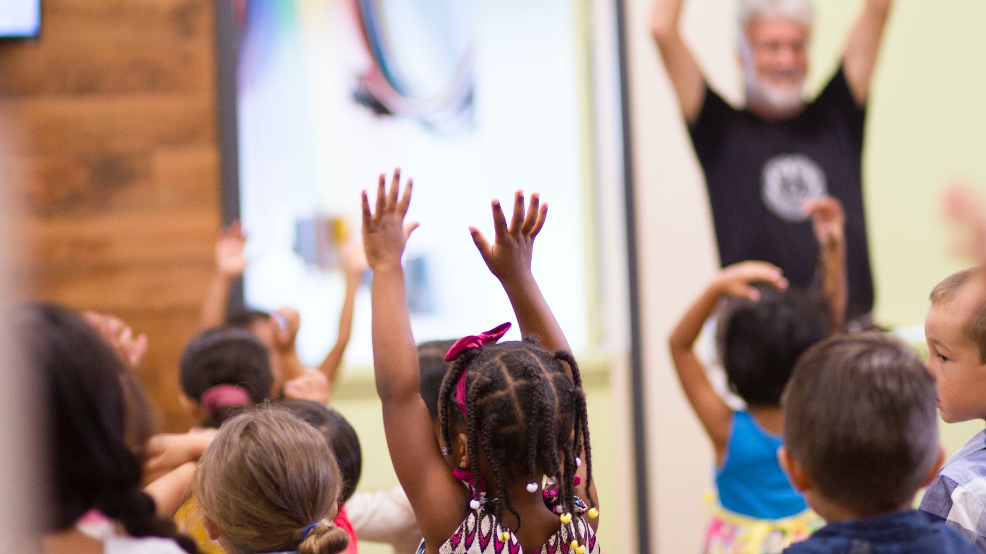 A child puts arms in the air in dance among a group of other children. An adults leads in the background.