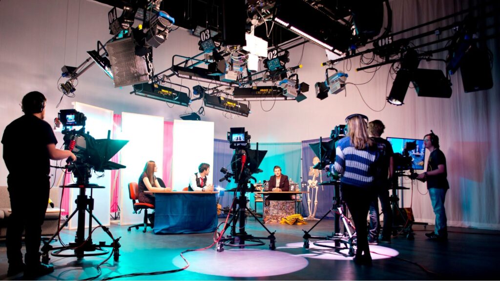 Students operate TV cameras, filming presenters in a studio, with a lighting rig overhead.