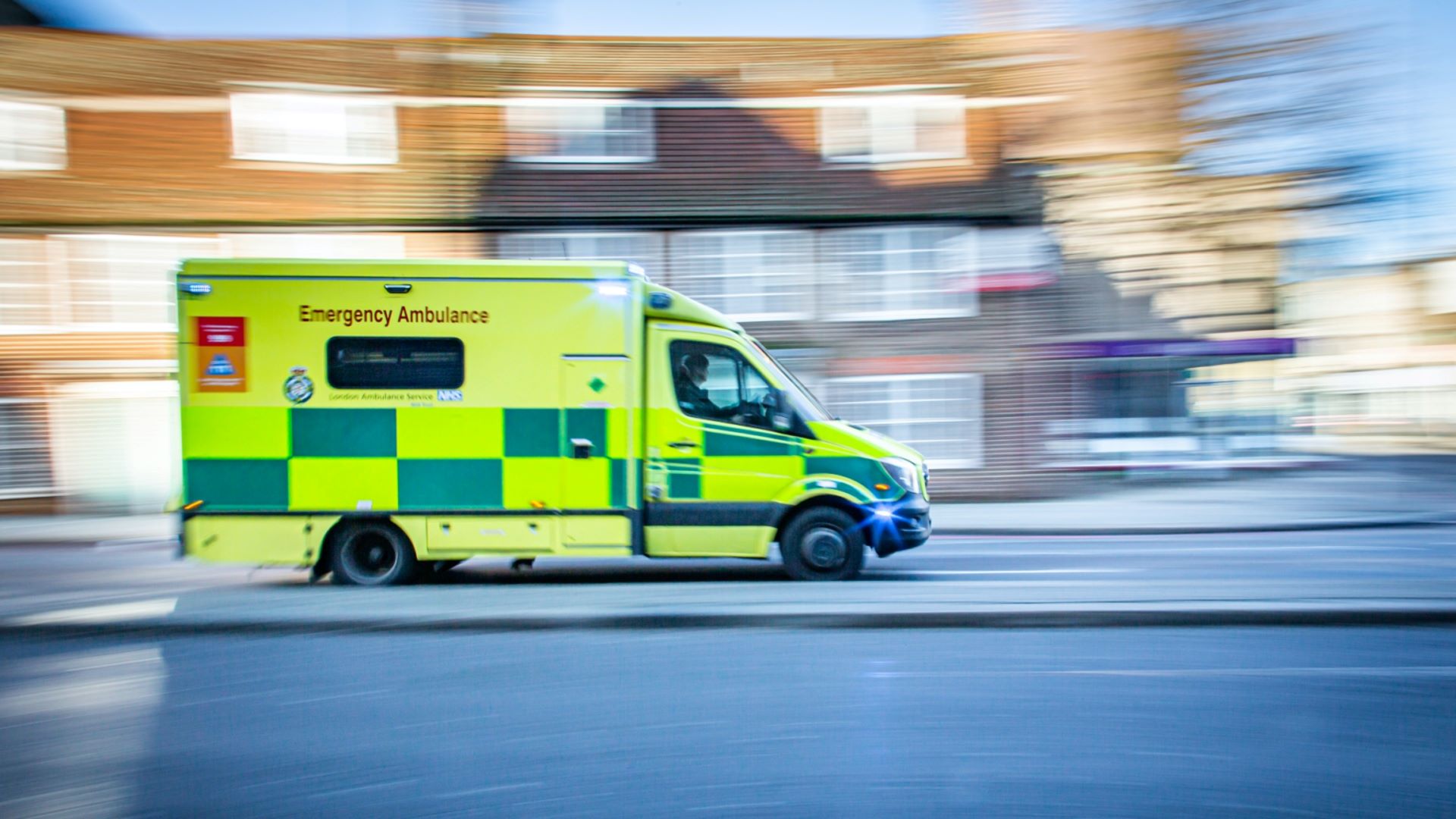 An image of an ambulance being driven on a road with some houses blurred in the background.