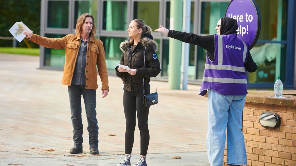 A campus connector holding their 'Here to Help' Lollipop and wearing their purple edge hill university vest gives people directions