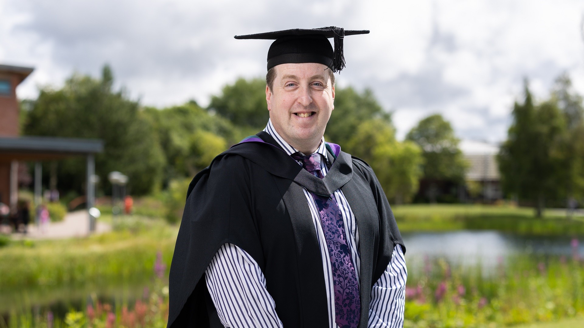 Andrew Sutcliffe wears a cap and gown and stands in front of a lake.