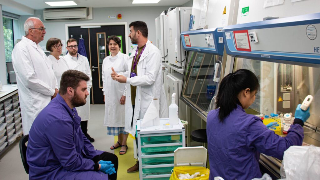 A picture with five people in white lab coats and two people in purple lab coats. Those in white are watching the other two conduct experiments using lab equipment.