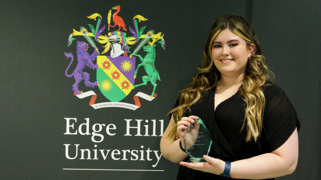 Student Élise Westbury-Jones posing with her glass award in front of the Edge Hill University crest.