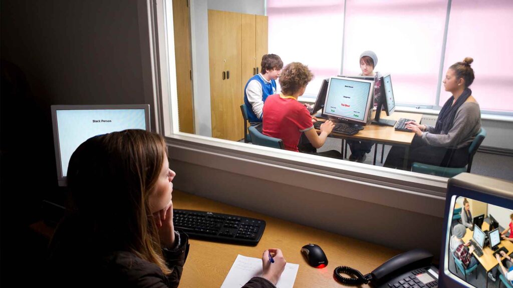 A group of students taking part in a Psychology study. A teacher is behind the glass and four students are looking at computers at a table.