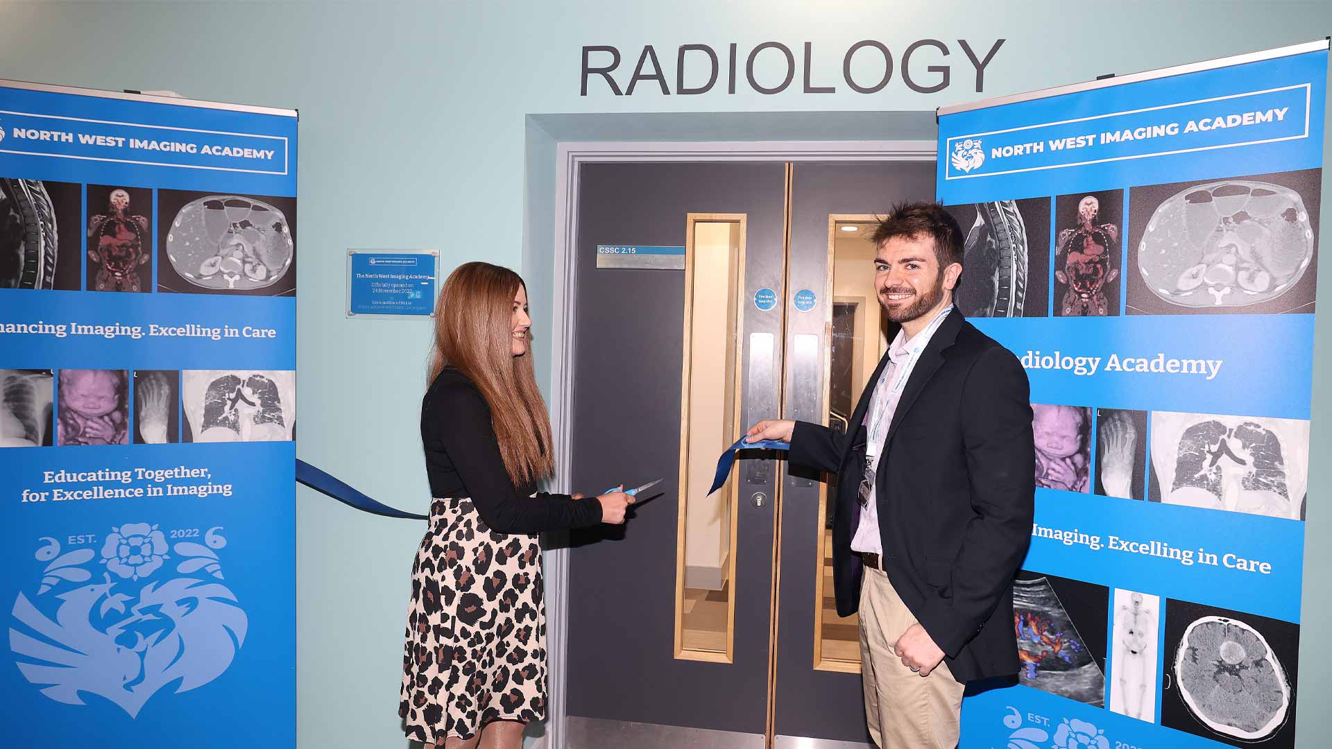 Two people stand outside the doors of a radiology department, cutting a ribbon to open it. 