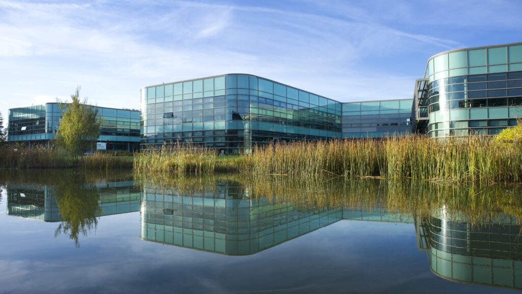 A glass and metal building is reflected in the surface of a lake with blue sky overhead.