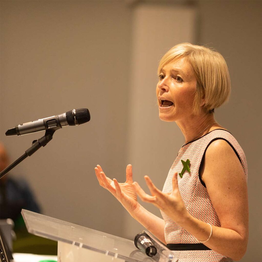 Jo Crotty on stage (a woman with a blonde bob and cream dress talking into a microphone).