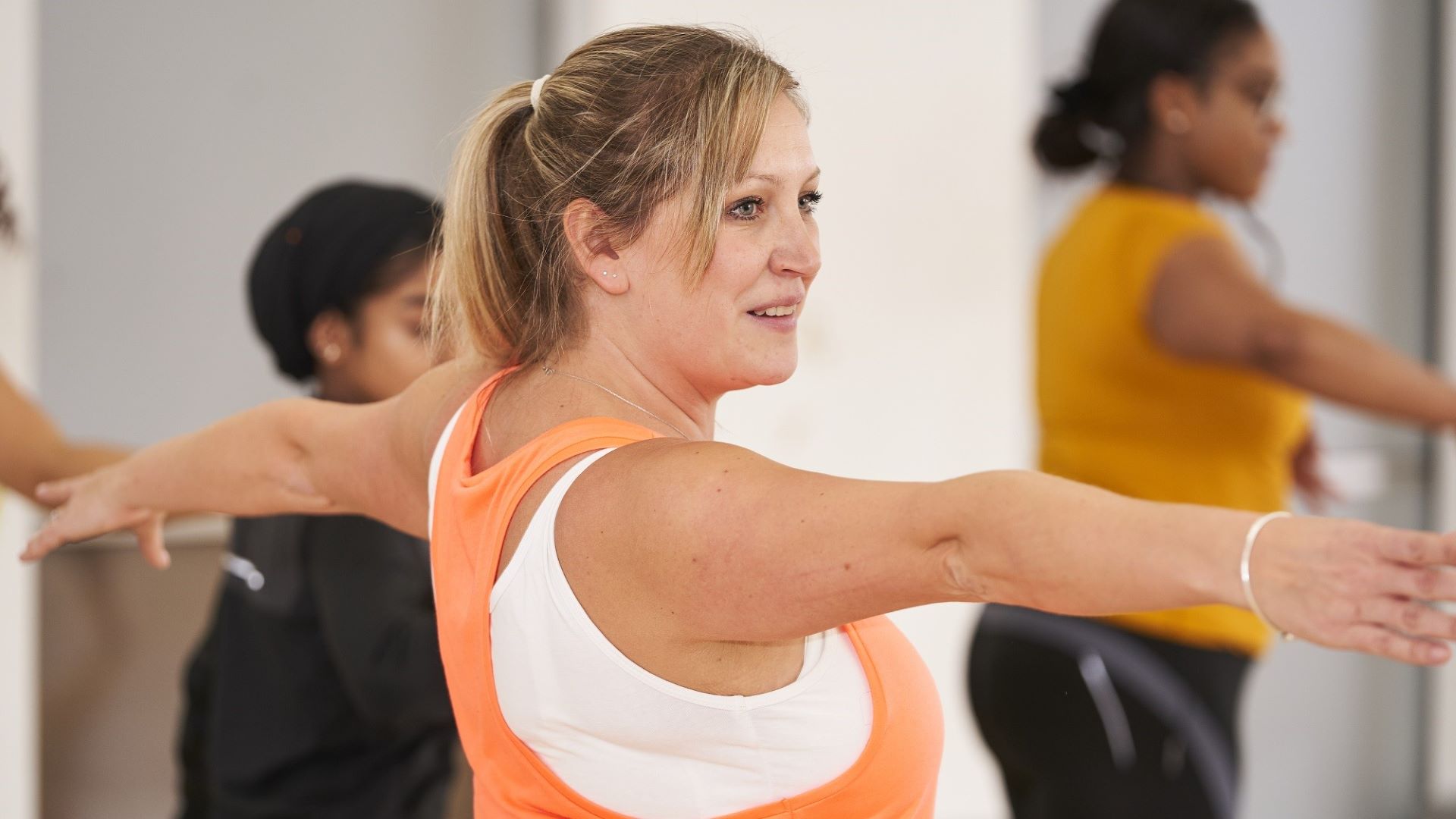 A woman wearing an orange sports top stretches her arms out in a physical activity class.