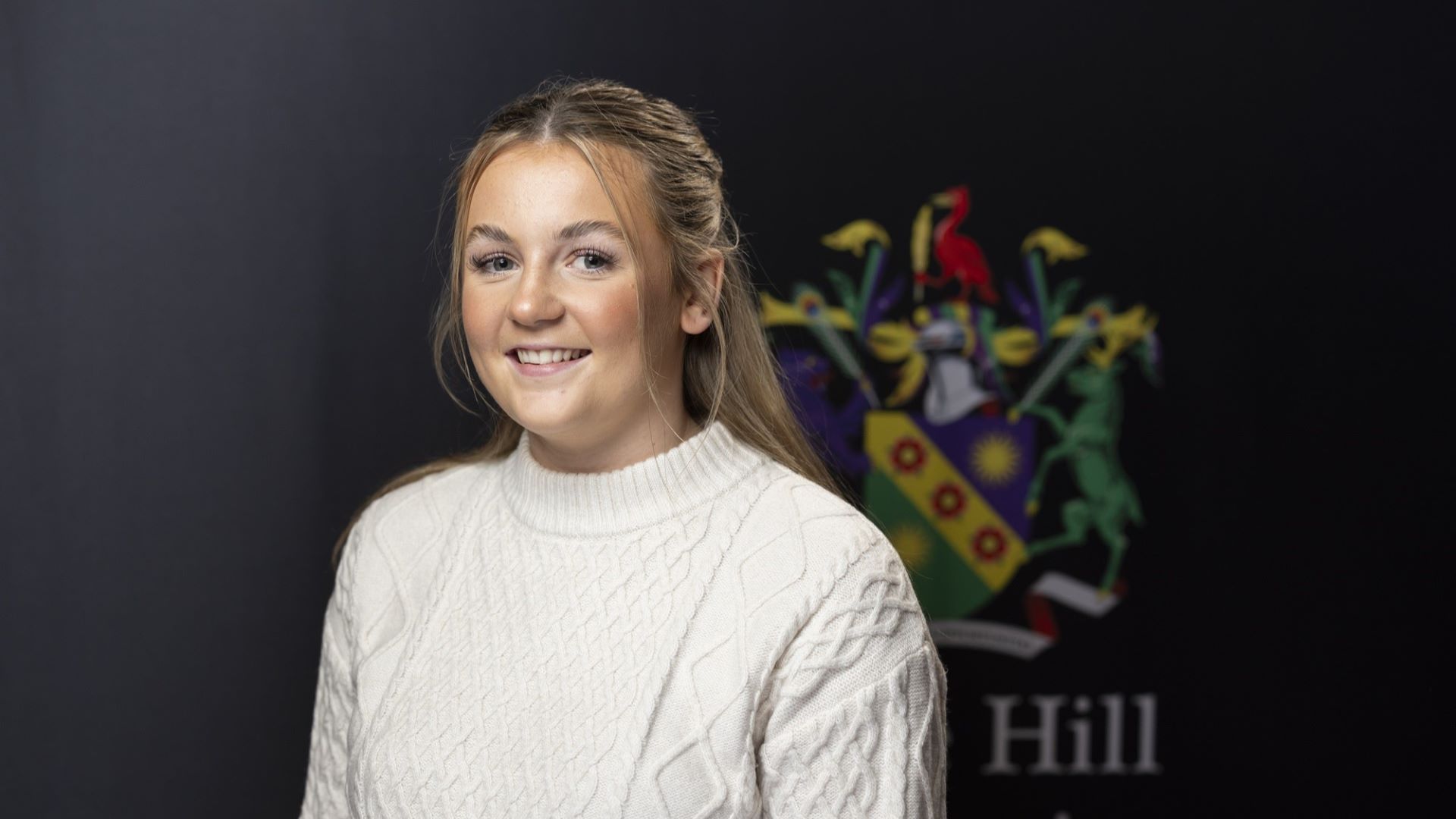 Student Grace Arrowsmith smiles at the camera while standing in front of the Edge Hill University crest