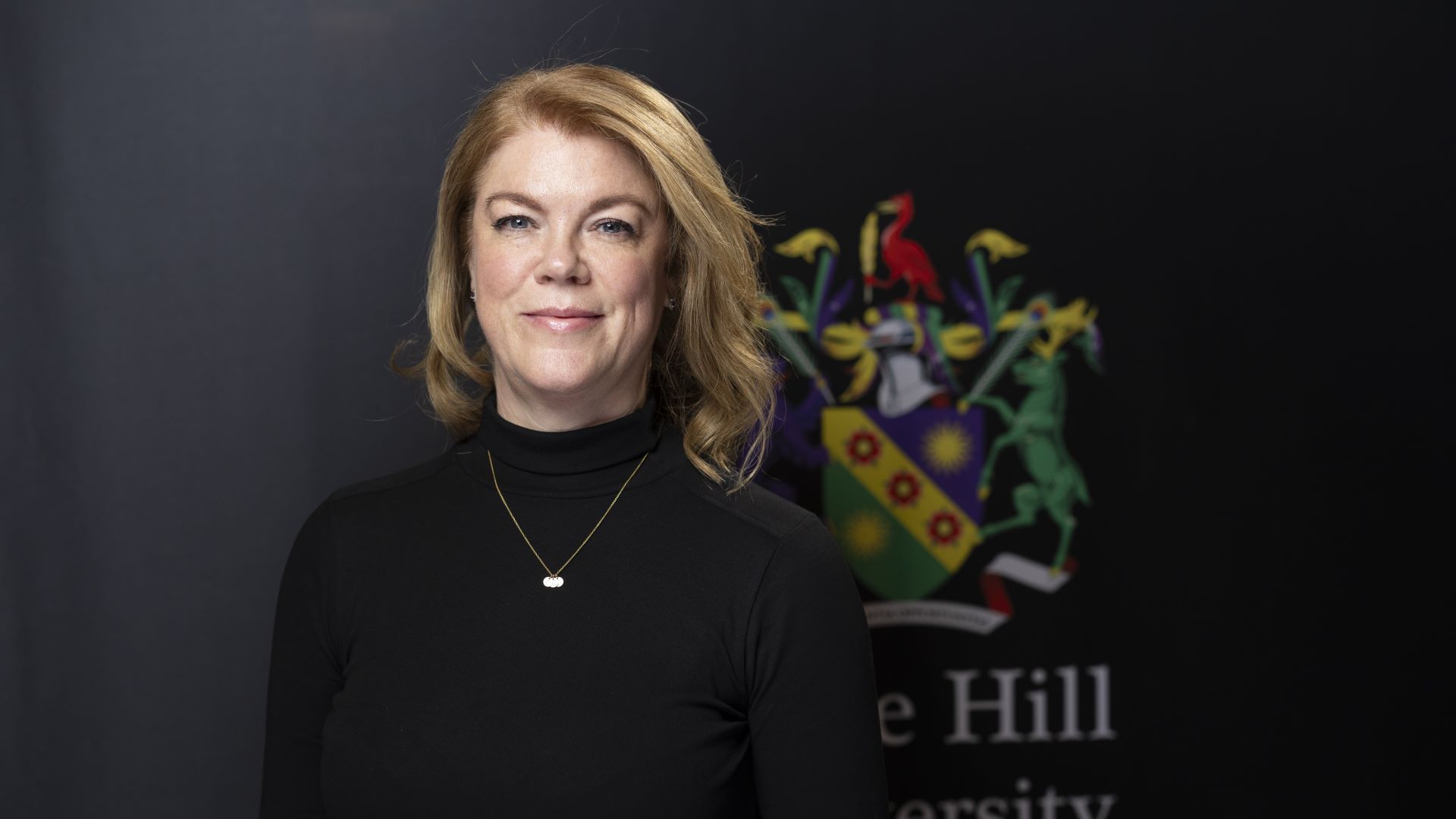 Jo Woodward standing in front of Edge Hill University crest.