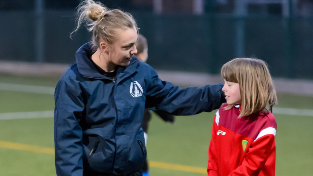 A sports coach talks to a child on a pitch