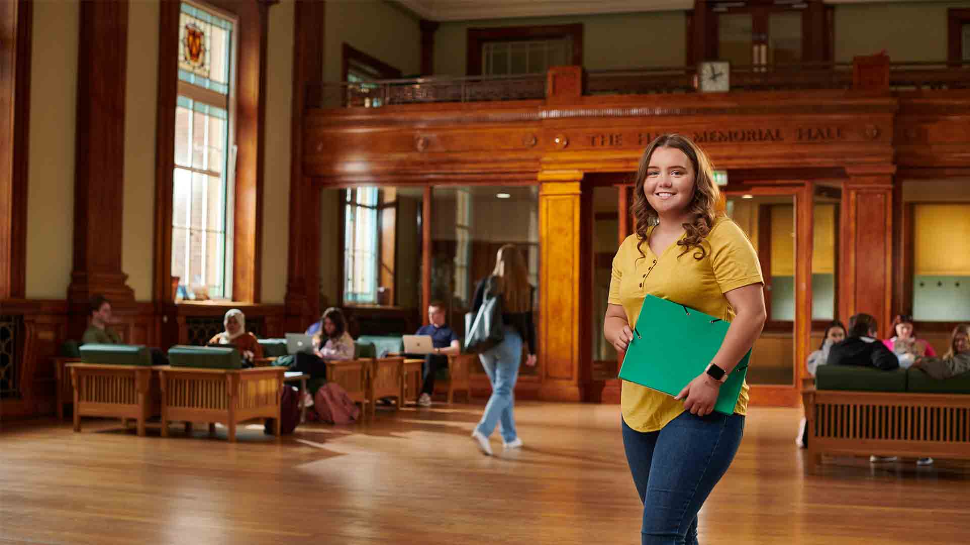 Female student stood in Hale Hall holding a green folder.
