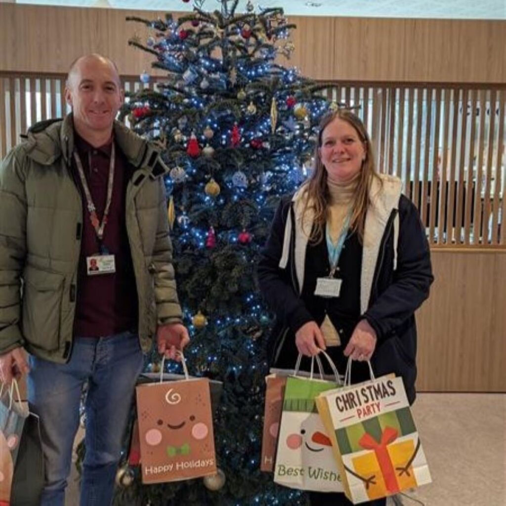Two people stand in front of a Christmas tree with gift bags in their hands.