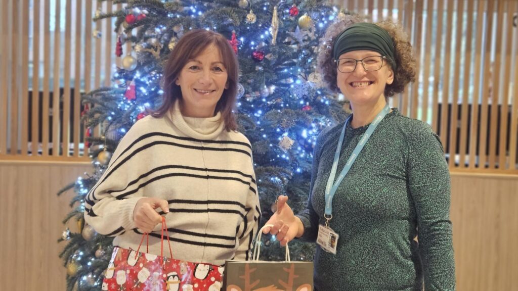 Helen Rimmer hands Christmas gift bags to Karen Cooke in front of a large decorated Christmas tree.