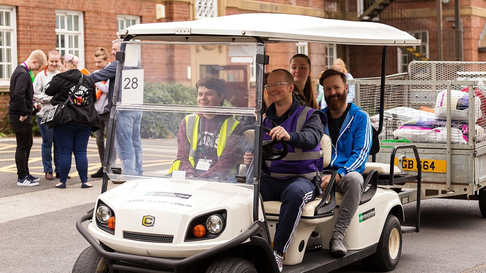 A member of Edge Hill staff drives a golf buggy with three visitors and their luggage on board.