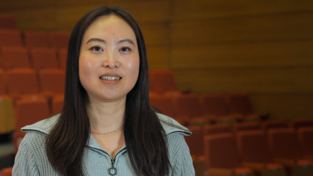 Dr Fei Chen sits in a lecture theatre and smiles at the camera, discussing the children's mental health project, Arts4Us.