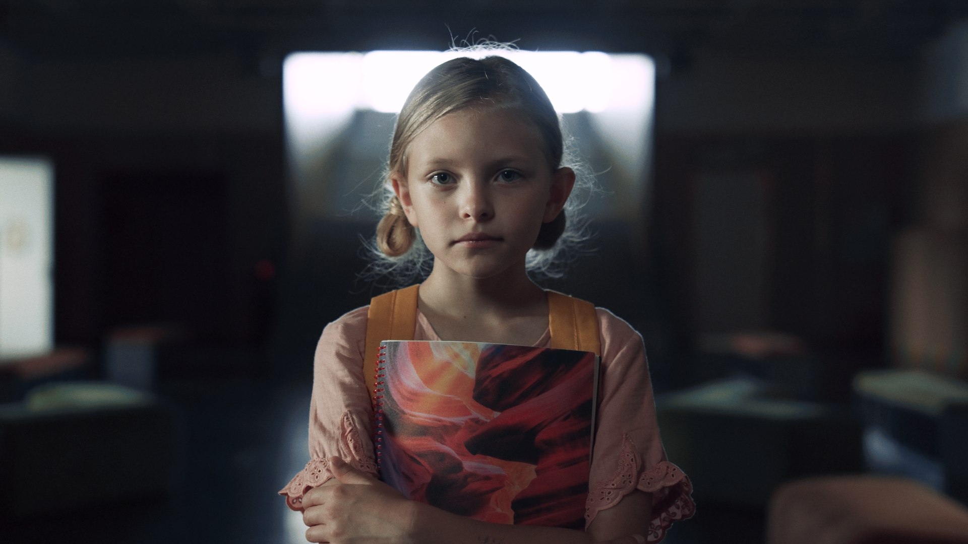 A young girl looks sadly at the camera while clutching a book to her chest.