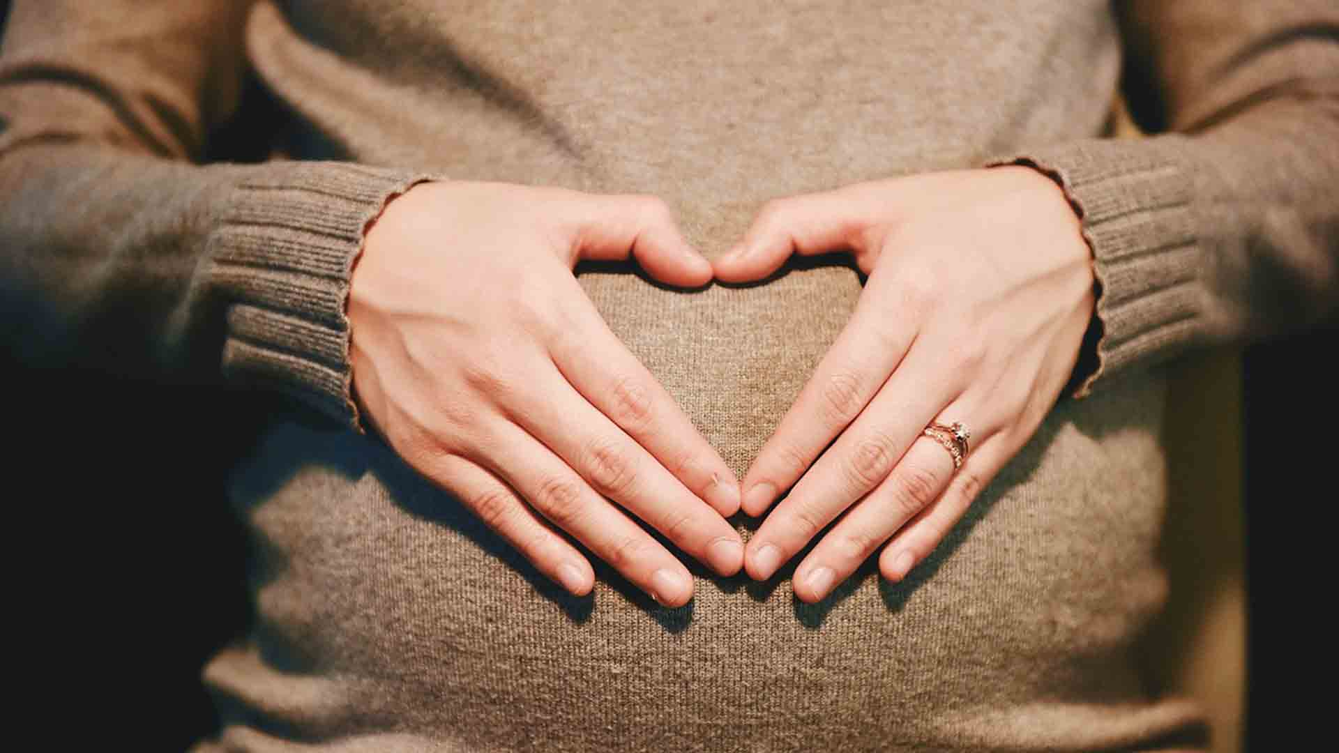 A pregnant woman holds her hands on her stomach in the shape of a heart.