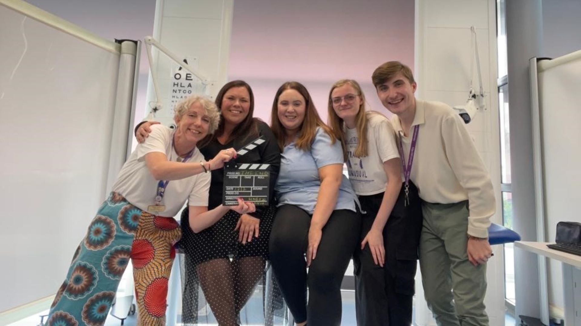 A group of students and staff gather together on a medical bed. One holds a directors clapperboard.