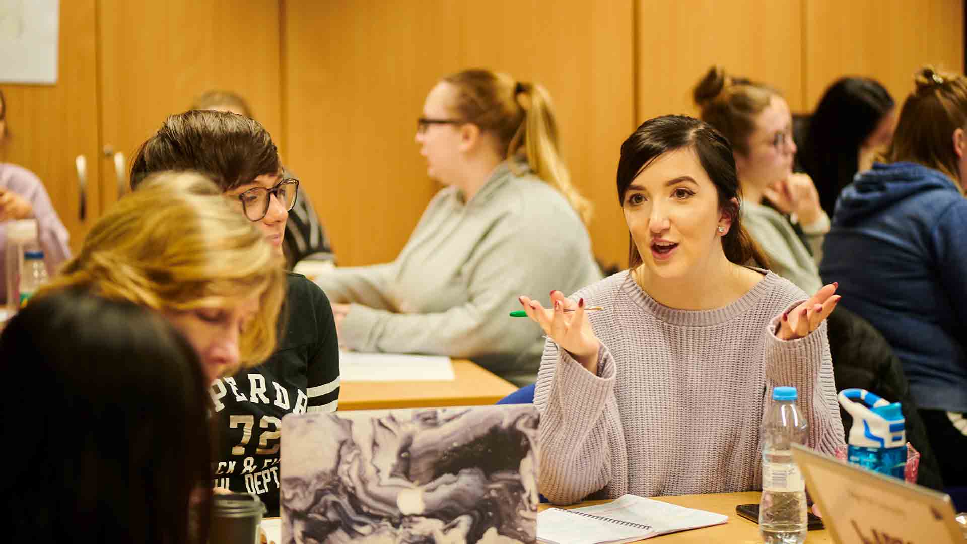 Women sat a wooden table in a busy room. One woman has her back to the camera, one is obscured from view, and one is mid-talking, wearing a purple jumper with dark hair.