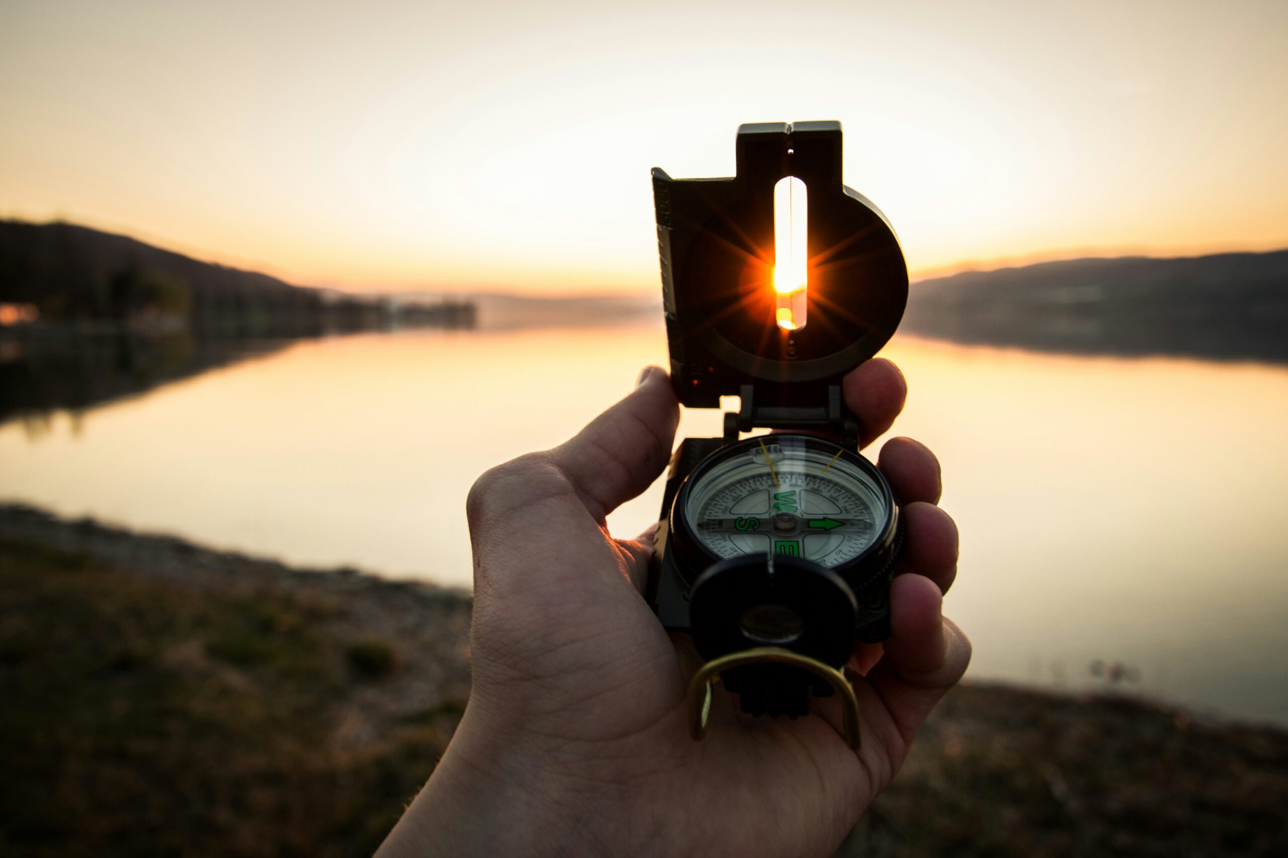 A photo of someone's hand holding a compass with a backdrop of a lake at sunset to represent navigation