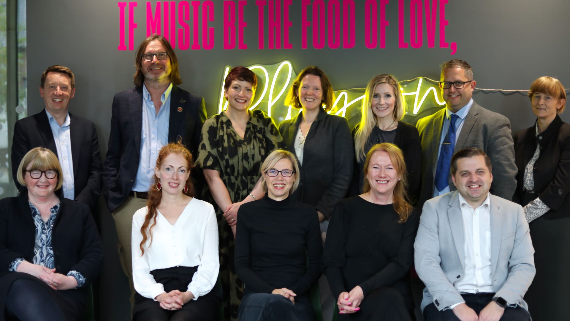 12 people stand and sit in front of a neon sign saying "If music be the food of love, play on".