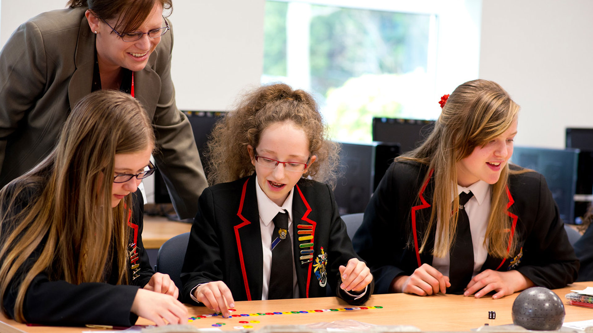 Three girls sat at a desk in a secondary school working with dice and counters. A teacher stands behind them encouraging them.