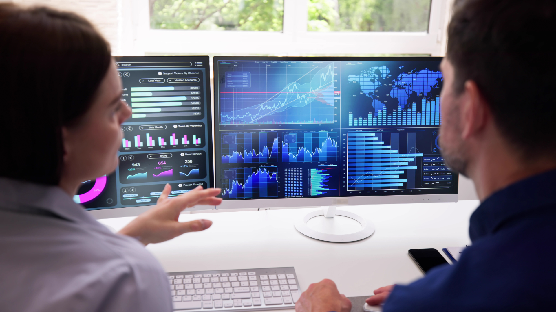 Two people look towards two computer screens which display data.