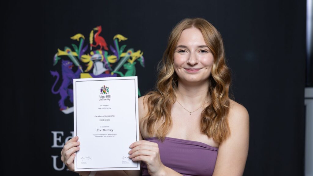 Zoe Harvey stands smiling with her certificate in front of an Edge Hill University logo.
