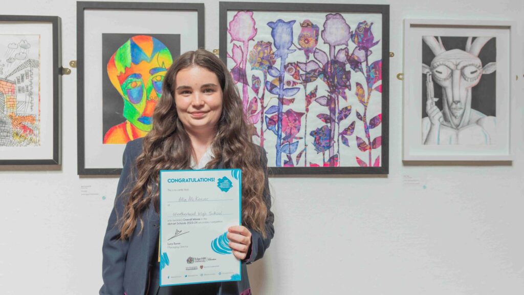 A young student dressed in grey school uniform, standing in front of 4 pieces of art work, holding a winner certificate.
