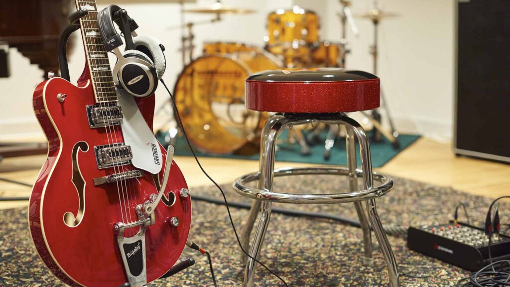 Guitar on a stand, with a drum stool, and drums in the background.
