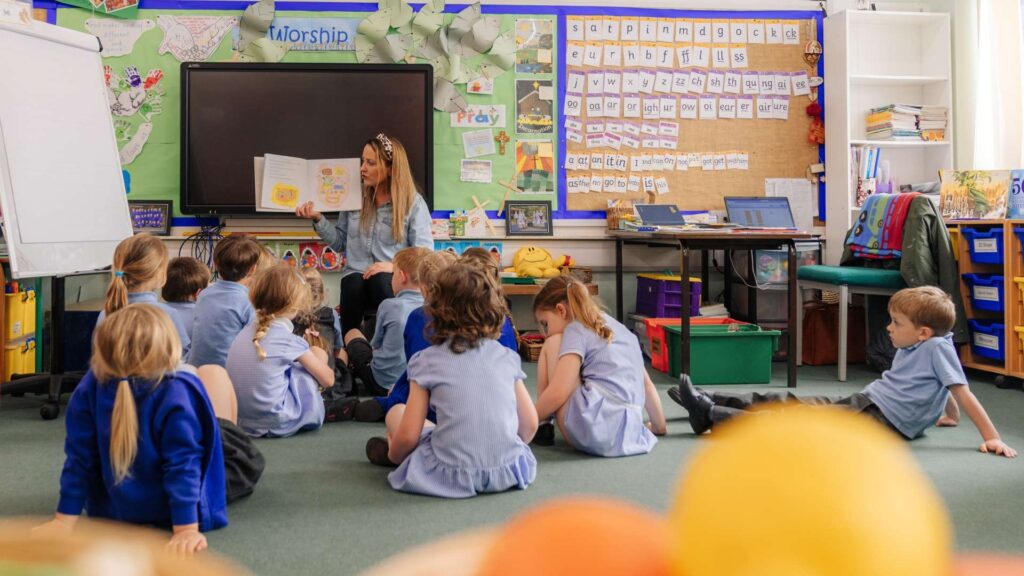 a primary shcool class room with students wearing blue uniform as the teacher reads a book to them.