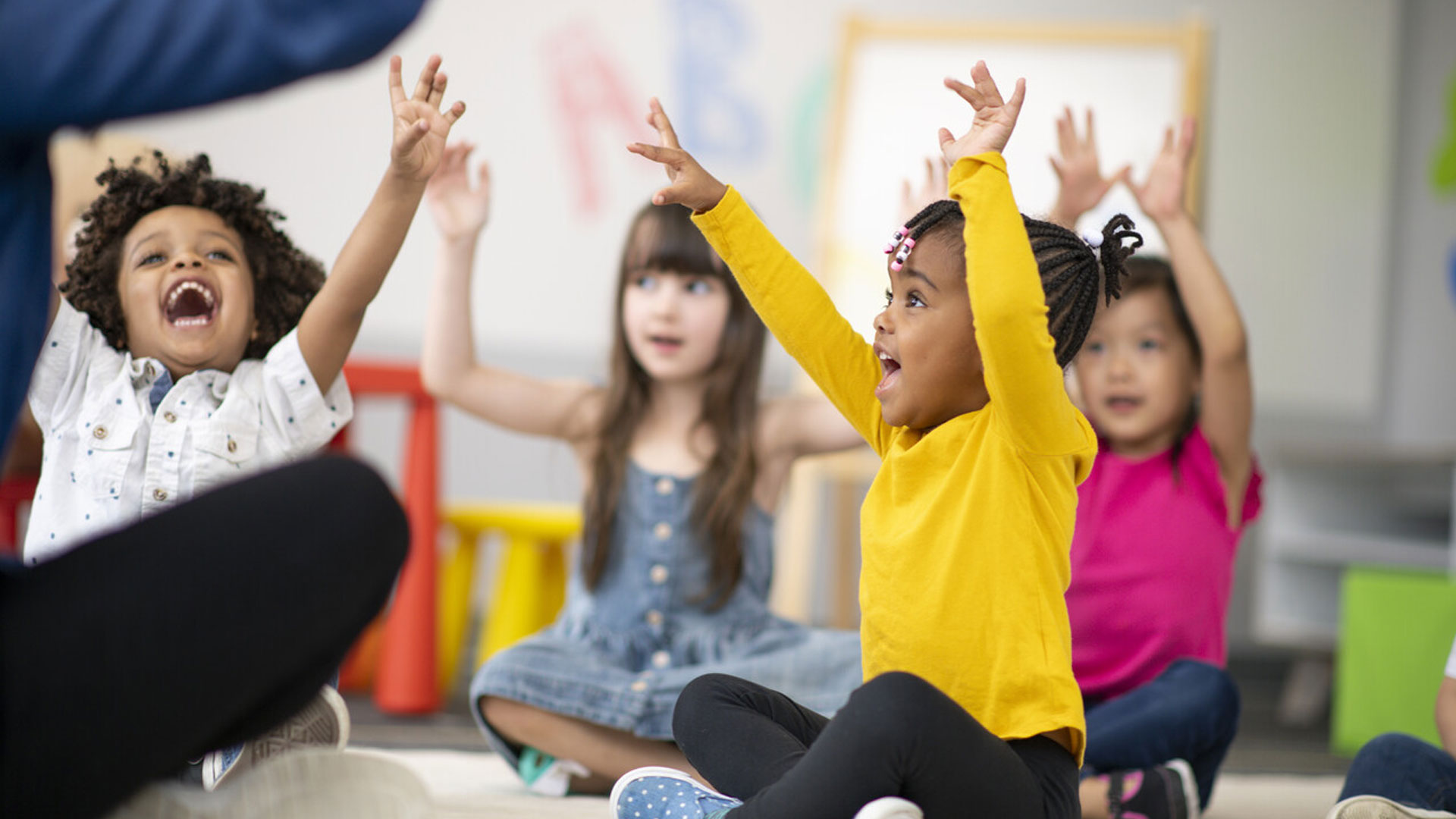 A group of young children raise both their arms in the air in a classroom