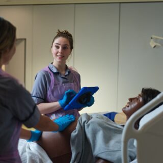 Image of Nicole Lightfoot in the midwifery ward looking towards another student whilst holding a tablet device at the bedside of a birthing mannequin.