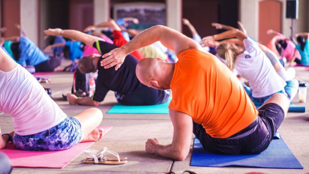 Several people in colourful gym wear sit on mats and stretch their right arms over their heads