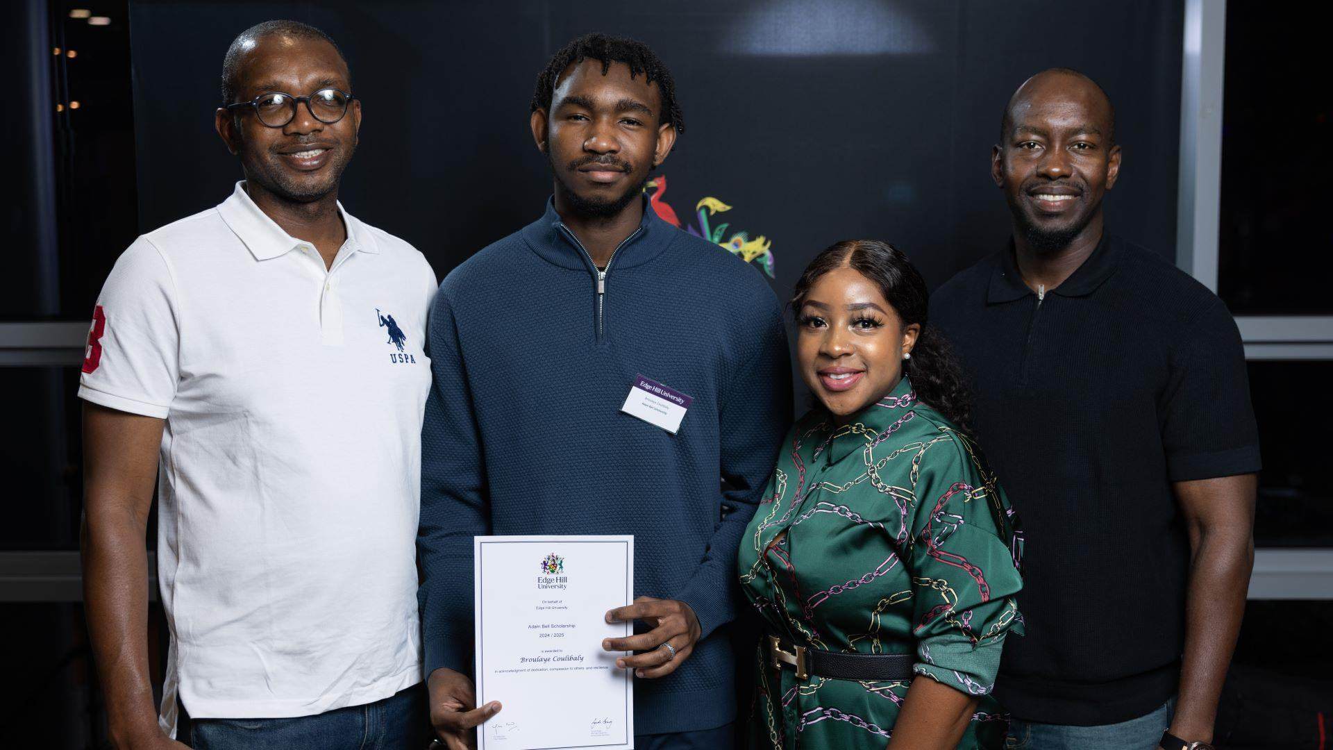 Broulaye and three supporters stand together in front of an Edge Hill University logo. Broulaye holds a certificate.