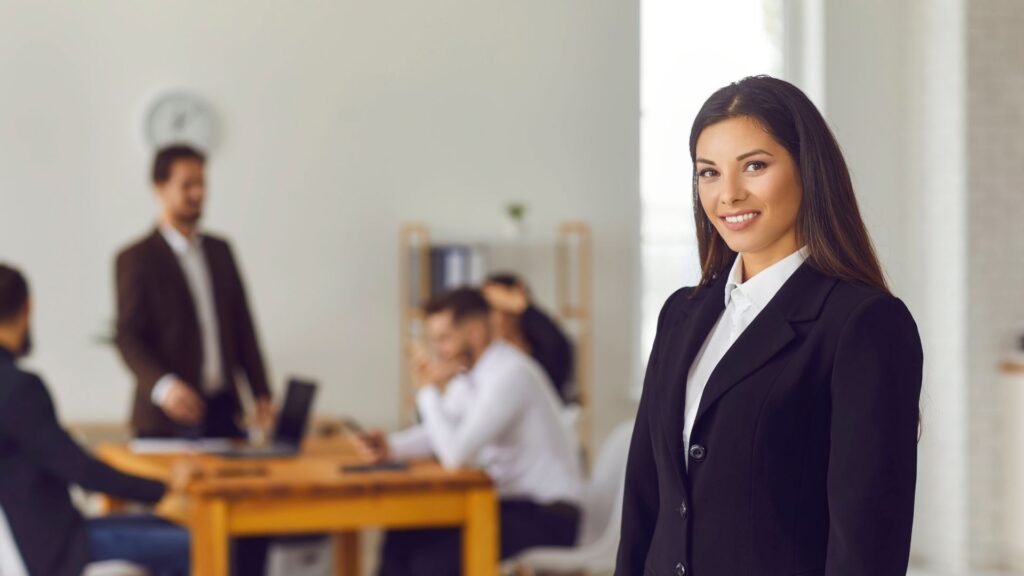 A business woman in the foerfront of the image with a team behind her of men sat at a desk. 